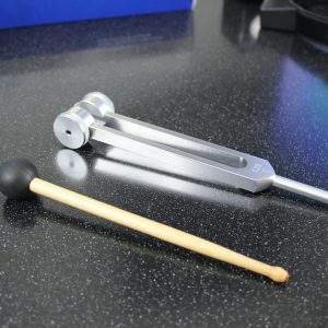 A shiny metal tuning fork with mallet on speckled black table, for sound therapy or music.